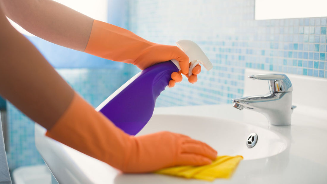 Person mit orangefarbenen Handschuhen reinigt Waschbecken mit Tuch und Sprühflasche, Nahaufnahme im Badezimmer