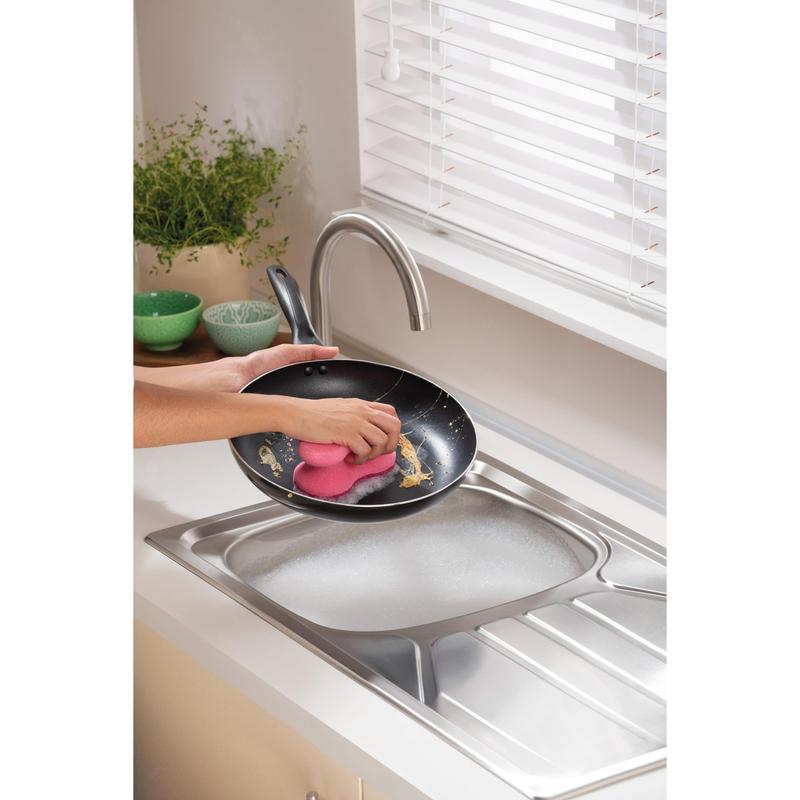 A person cleans a frying pan under running water in a modern kitchen with a 3M Deutschland GmbH Scotch-Brite® Delicate Color Comfort non-scratch cleaning sponge (2 pieces) - ideal for delicate surfaces. Green bowls and a plant are on the countertop.