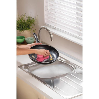 A person cleans a frying pan under running water in a modern kitchen with a 3M Deutschland GmbH Scotch-Brite® Delicate Color Comfort non-scratch cleaning sponge (2 pieces) - ideal for delicate surfaces. Green bowls and a plant are on the countertop.