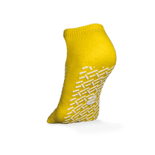 A single yellow ECO-PLUS anti-slip disposable sock, double-sided coated by AMPri Handelsgesellschaft mbH, stands upright on a white background, with white geometric anti-slip grips and a central logo, ideal for medical areas.