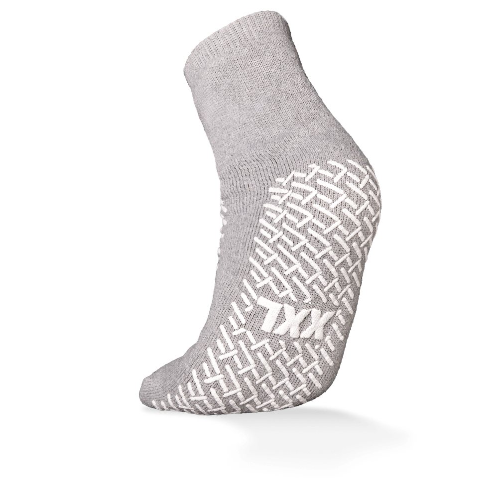 Pictured is a single gray ECO-PLUS anti-slip disposable sock from AMPri Handelsgesellschaft mbH with a white anti-slip grip and "TXX" lettering on the sole. Double-sided coated, ideal for medical areas; the background is plain white.