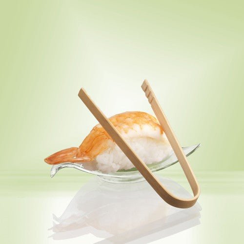 A pair of 10 cm PAPSTAR bamboo finger food tongs from PAPSTAR GmbH rests on a glass dish next to shrimp nigiri - ideal for serving snacks or party finger food - against a background with a pale green gradient.