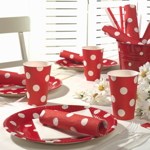 A table is set with red paper plates, cups, and 20 PAPSTAR napkins (40x40 cm, 3-ply) from PAPSTAR GmbH, all with white dots. White daisies adorn the table while light falls through white blinds in the background.