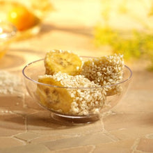 Three pieces of banana topped with white sesame seeds are served in a Starpak 50 finger food tray (PS crystal clear), placed on a light-colored tiled surface with a softly blurred background—ideal for events.