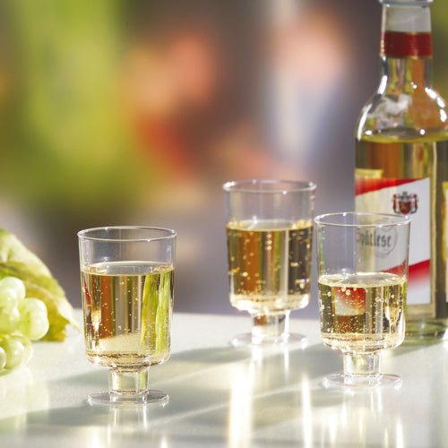 Three Starpak stem glasses for white wine (PS, 0.1l, crystal clear) are filled and stand next to a bottle of white wine and green grapes on a white surface in front of a blurred background.