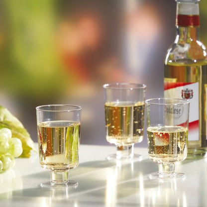 Three Starpak stem glasses for white wine (PS, 0.1l, crystal clear) are filled and stand next to a bottle of white wine and green grapes on a white surface in front of a blurred background.