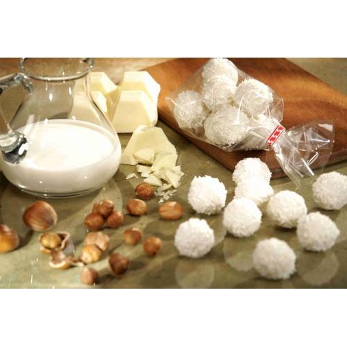 On a countertop, there is a glass jug with milk, white chocolate chips, hazelnuts, and coconut-covered candy balls arranged, with some treats presented in a Starpak 300 flat-bottom bag (transparent PP) from Starpak on a wooden cutting board.