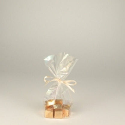 A Starpak 10 bottom bag, PP transparent from Starpak, tied with a cream-colored ribbon and filled with gold-wrapped square candies, stands on a simple, light surface.