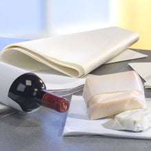 A wine bottle stands on a gray surface, next to PAPSTAR parchment substitute "pure" white wrapping paper (12.5 kg, grease-resistant from PAPSTAR GmbH) and two blocks of cheese, one of which is partially wrapped. The background is slightly blurred.