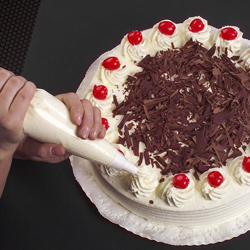 A pastry chef decorates a round cake with whipped cream using the Starpak 100 transparent piping bag from Starpak; the cake, garnished with chocolate shavings and cherries, lies on a white paper doily on a dark background.