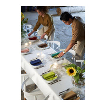 Two people arrange colorful napkins on a long table set with glasses and cutlery. A Dunicel tablecloth roll (1.18 m x 10 m) from Duni GmbH covers the table and adds the charm of large events to the inviting interior.