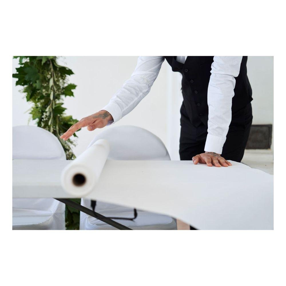 A person in a white shirt and black vest rolls out a Dunicel tablecloth roll from Duni GmbH (1.18 m x 10 m) over a table, with white chairs and green foliage visible in the background.