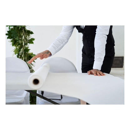 A person in a white shirt and black vest rolls out a Dunicel tablecloth roll from Duni GmbH (1.18 m x 10 m) over a table, with white chairs and green foliage visible in the background.