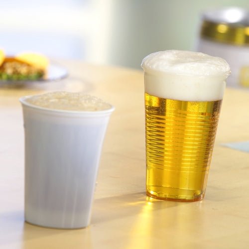 Two beer cups are on a light wooden table - one in a clear Starpak 75 drinking cup, PS clear with foam rim from Starpak, showing golden beer, the other in an opaque white cup. Blurred food can be seen in the background.