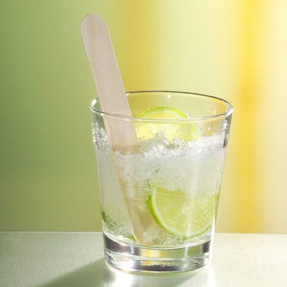 A glass with crushed ice, lime slices, and a PAPSTAR wooden drink stirrer 15 cm (ideal for Caipirinha cocktails) from PAPSTAR GmbH stands on a reflective surface in front of a blurred yellow and green background.