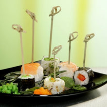A black plate is presented with various types of sushi, each skewered with a PAPSTAR 250 Fingerfood-Picker "pure" of PAPSTAR GmbH. The whole is garnished with roe, leafy greens, and wasabi; the background is in soft green and yellow tones.