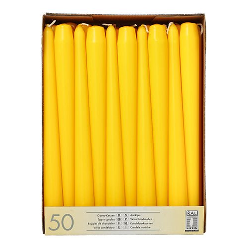 A cardboard box from Starpak contains 50 white taper candles (Ø 2.2 cm - 25 cm), arranged upright in rows. The label on the front contains product information in multiple languages, including "paraffin candles."