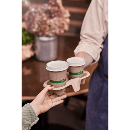 A person hands over a paper tray with two 120 ml Melli cups with white lids and green bands from Duni GmbH, proudly displaying their FSC™ certification. In the background, flowers and a wooden table can be seen, conveying an eco-friendly atmosphere.