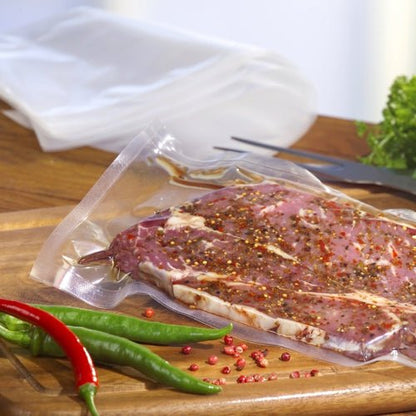 A Starpak 100 vacuum seal bag, PA/PE transparent from Starpak holds marinated raw meat on a wooden board with green and red chili peppers, tongs, and leafy greens - ideal for your chamber vacuum sealer.