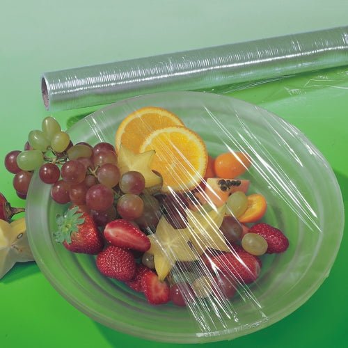 A transparent sheet of Starpak perforated cling film covers a bowl with various fruits, while behind it on a green surface lies a roll of unused Starpak perforated cling film.