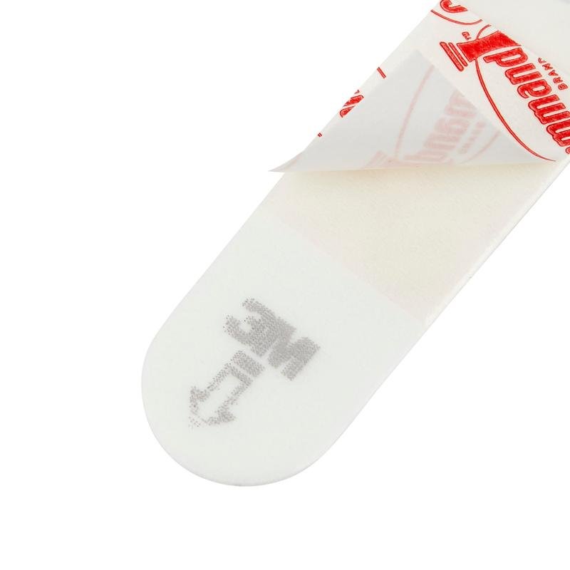 A close-up of a Kleine Command™ Strips S, 17022N from 3M Deutschland GmbH shows its rectangular shape, with one end of the protective paper peeled off, revealing the adhesive. "MC" and an arrow are printed to allow for easy and damage-free use on the wall.