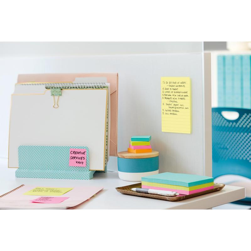 A tidy office desk with file folders, a notepad, colorful sticky notes, a pen, a tablet, and a round container. On the partition wall lies a yellow Post-it® Note in large format (101 mm x 152 mm) from 3M Deutschland GmbH, with blue chairs and cabinets in the background.