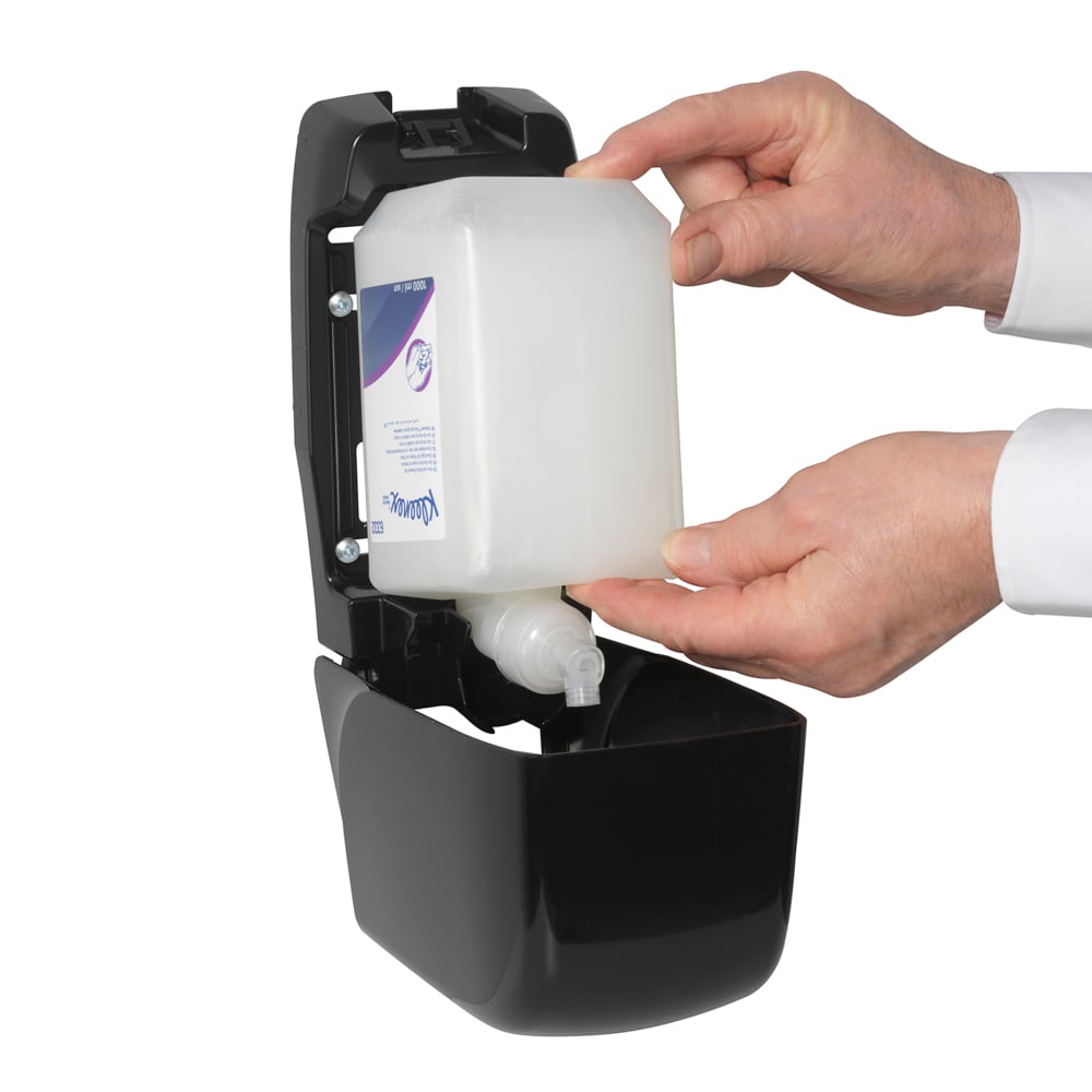 A person inserts a Kleenex® Hair and Body Shampoo 1L cassette from Kimberly-Clark GmbH into an open black wall dispenser, using both hands to ensure gentle care and effective cleaning.