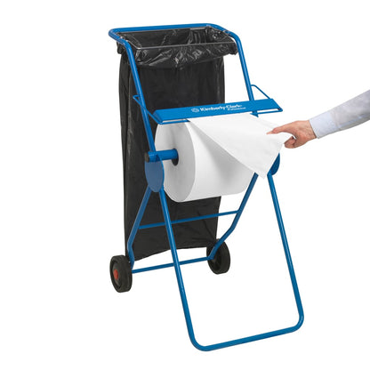 A blue paper towel dispenser with a large white Scott® ESSENTIAL™ medical roll (6 x 200 sheets) from Kimberly-Clark GmbH emphasizes hygiene; a black trash bag is nearby, while a person's hand pulls a sheet from the roll.