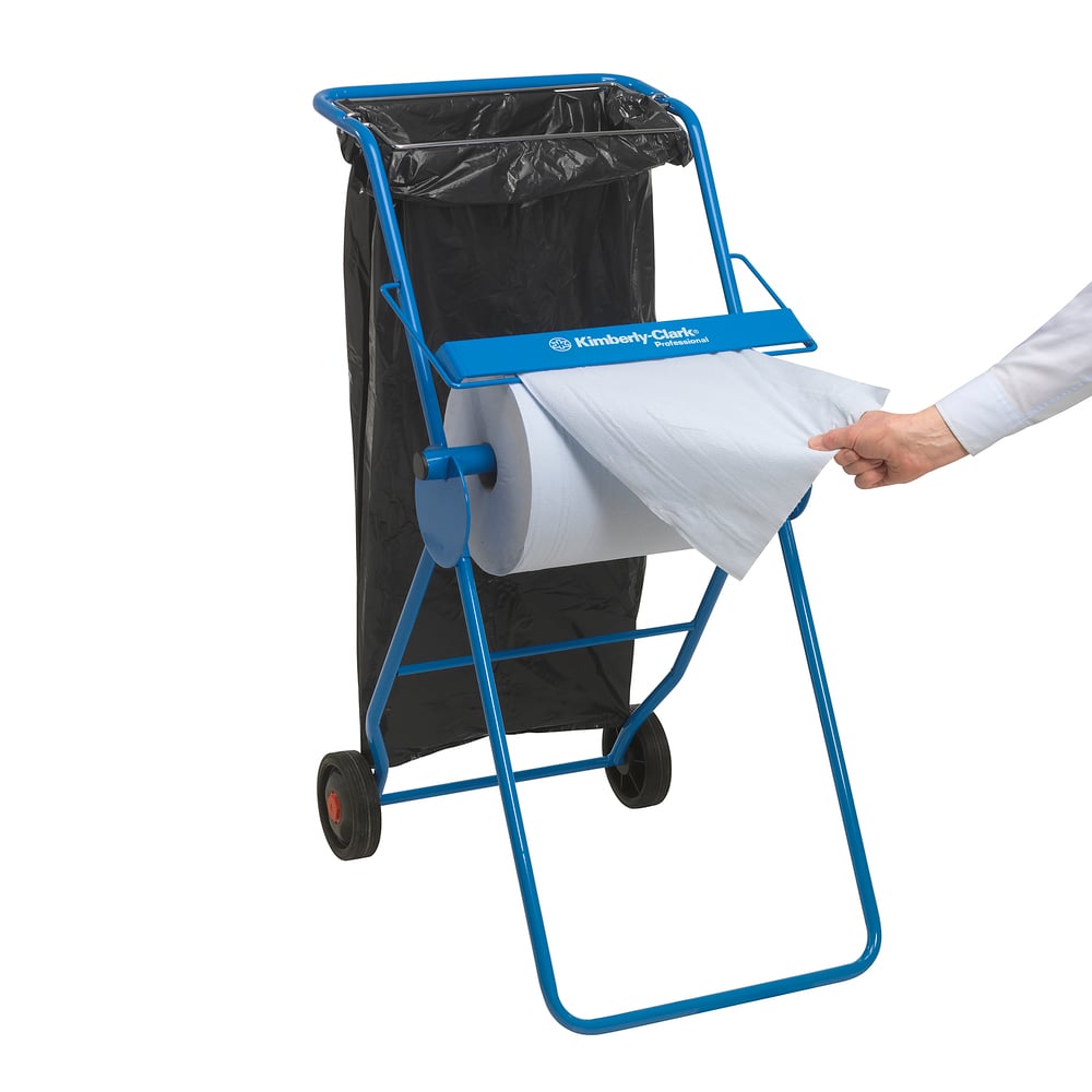 A person pulls a WypAll® L20 paper wipe for surfaces from a blue roll dispenser from Kimberly-Clark GmbH with two wheels, a metal frame, extra-wide white paper towels (1 roll x 1000 sheets), and a black garbage bag.