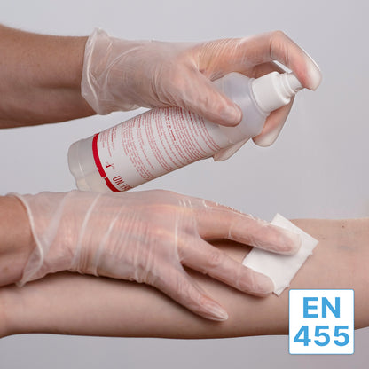 A person in ARNOMED VINYL PF tactile-sensitive disposable gloves from ARNOWA GmbH uses a spray bottle to moisten a cotton pad and clean another person's forearm. The label "EN 455" appears in the lower right corner.