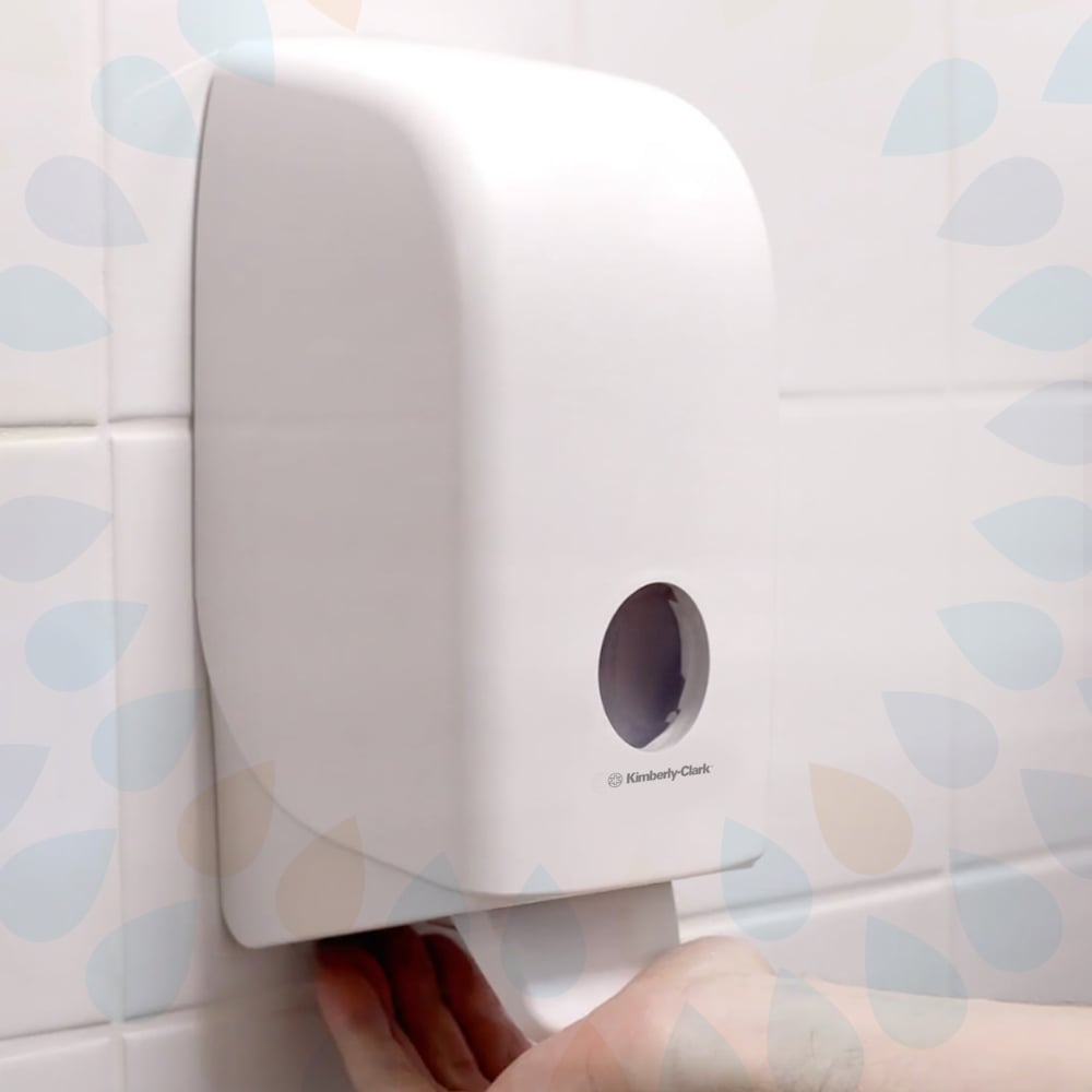 A hand is under a wall-mounted white paper towel dispenser in a tiled bathroom, next to a Kimberly-Clark GmbH Kleenex® Normal Wash Lotion Cassette Pink 1 Ltr (6 cartridges), with a paper towel being dispensed.