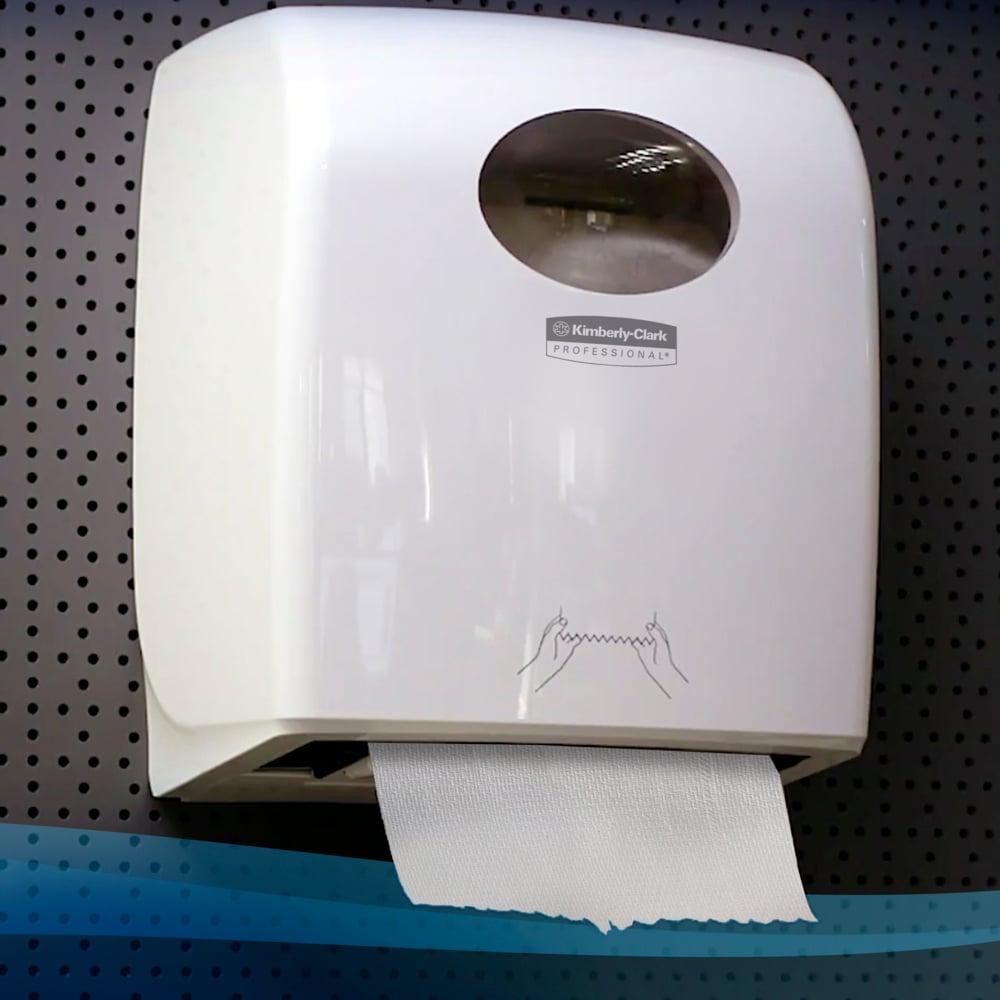 A white wall dispenser with a transparent viewing window holds a Scott® hand towel roll (304.00 m x 19.80 cm) from Kimberly-Clark GmbH with a hanging sheet on a perforated gray wall.