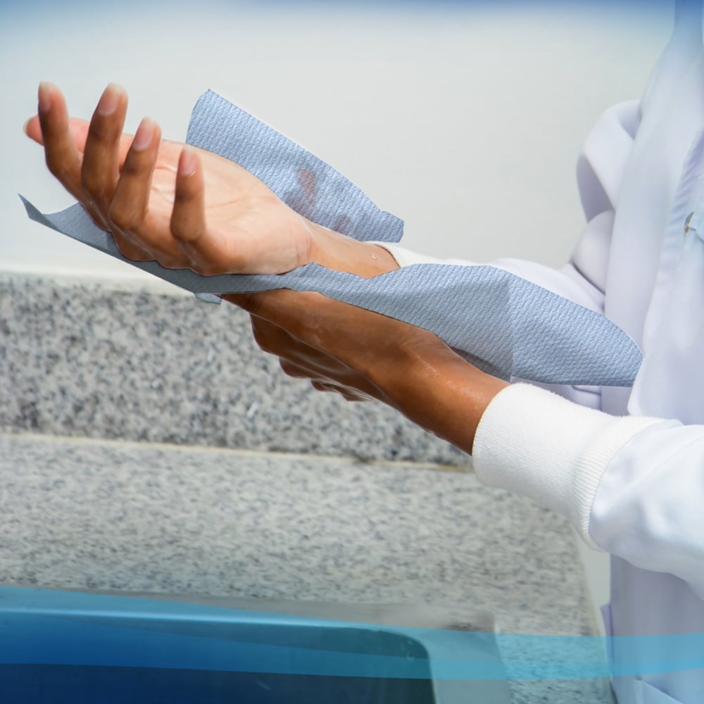 A person in a white coat dries their hands with Scott® hand towels - roll (304.00 m x 19.80 cm) from Kimberly-Clark GmbH from a paper towel roll next to a granite countertop and a sink.