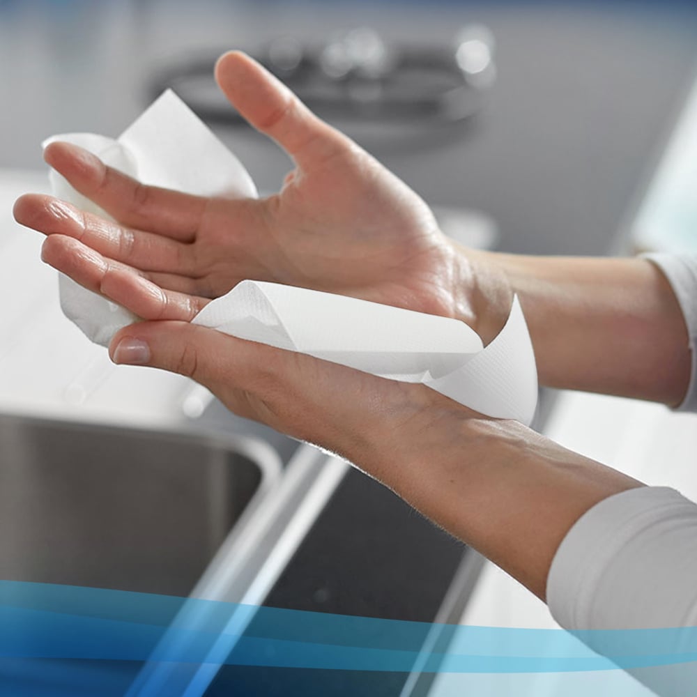 A person wipes their hands with a white Scott® hand towel roll (304.00 m x 19.80 cm) from Kimberly-Clark GmbH over a sink, with the countertop and sink visible in the background.
