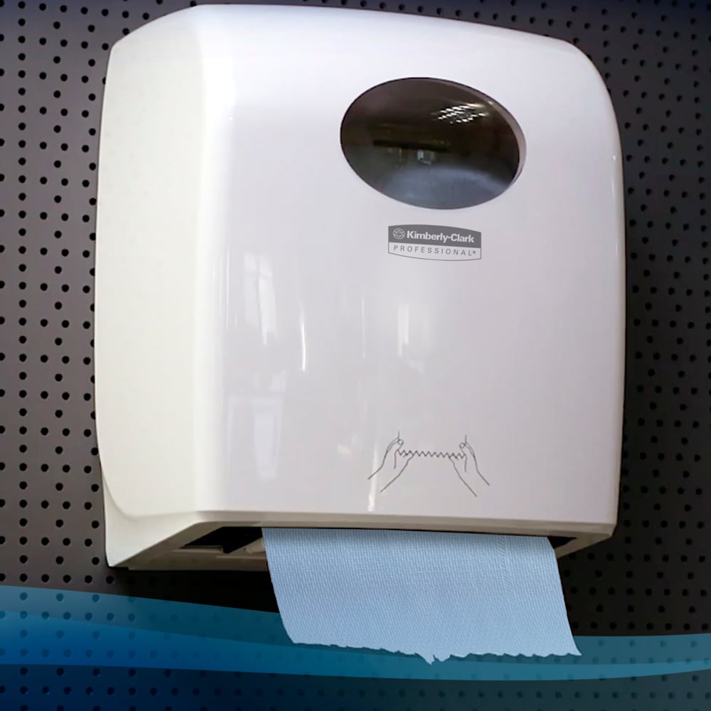 A white, wall-mounted dispenser from Kimberly-Clark GmbH holds a Scott® hand towel roll (304.00 m x 19.80 cm) with a blue towel and a simple, illustrated guide against a dark perforated metal background.