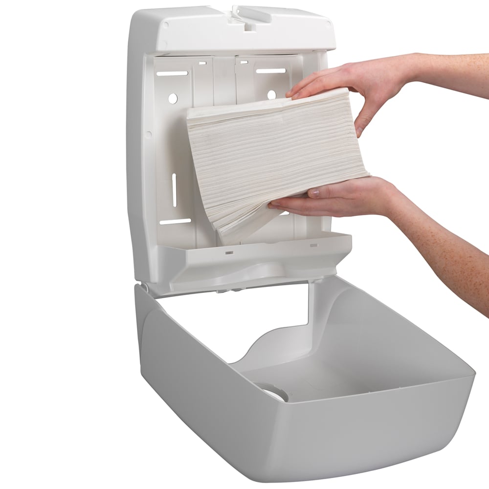 A person refills a wall-mounted dispenser with Kleenex® Ultra™ folded hand towels Interfold White (15 clips x 96 sheets) from Kimberly-Clark GmbH and ensures hygienic hand drying, as the open dispenser allows for easy refilling.
