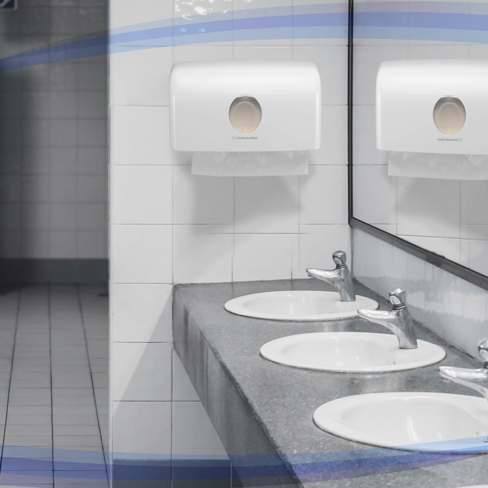 Three white sinks with silver fittings sit on a gray countertop. Above them, a mirror and the Kimberly-Clark GmbH Aquarius™ dispenser for folded towels (MultiFold/White/Small) provide hygienic towel dispensing on the tiled wall.