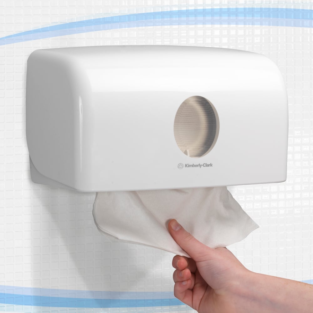 A hand takes a towel from the white Aquarius™ dispenser for folded towels (MultiFold, small) from Kimberly-Clark GmbH, which is wall-mounted on a tiled wall for hygienic dispensing.