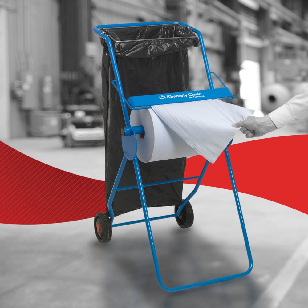 A blue Kimberly-Clark floor stand holds a WypAll® L20 large roll paper wipe for maintenance work and a black trash bag. A hand pulls a sheet - ideal for indoor industrial maintenance work.