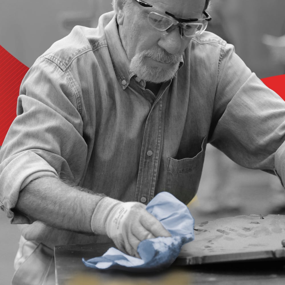 A person with safety glasses and gloves wipes a metal surface with a blue WypAll® L30 paper wipe from Kimberly-Clark GmbH. The gray-toned, blurred background features subtle red and yellow accents reminiscent of industrial cleaning tasks.