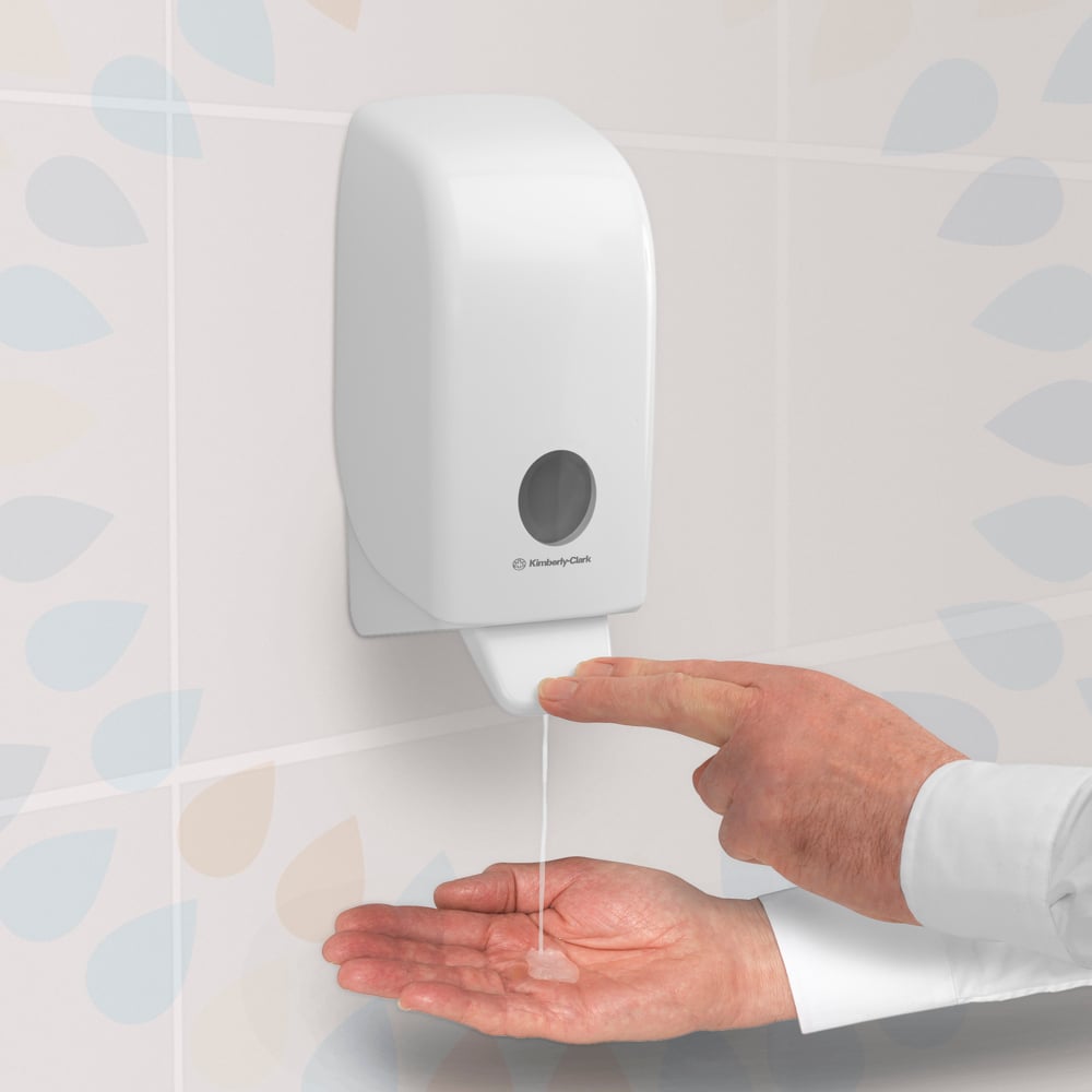 A person presses on a transparent Kleenex® Extra Gel Hand Sanitizer dispenser from Kimberly-Clark GmbH, mounted on a tiled wall, and dispenses the hand sanitizer gel from a 1-liter cartridge onto their open palm.