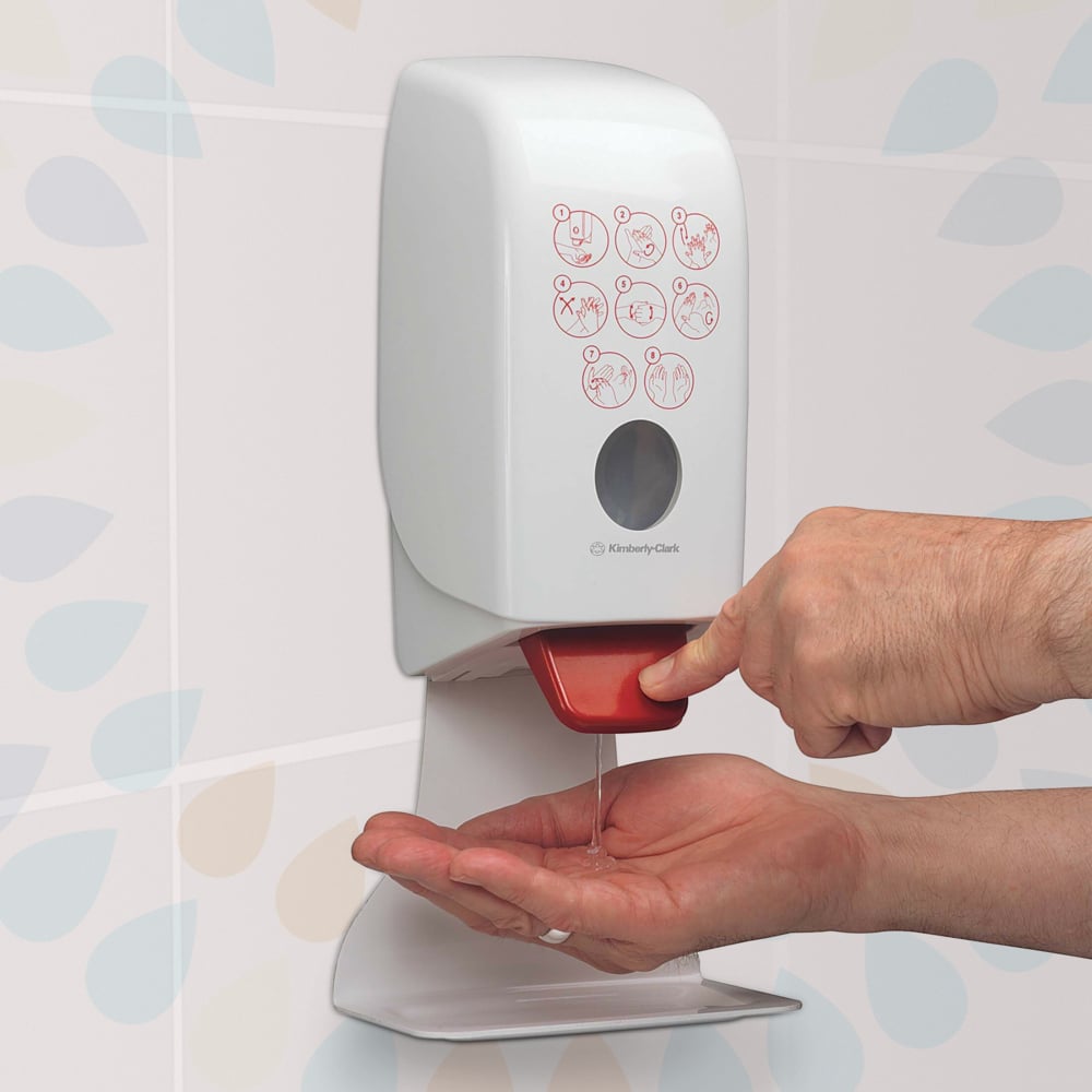 A person presses a red lever on a wall-mounted Kleenex® Extra Gel Hand Sanitizer dispenser (Kimberly-Clark GmbH) and dispenses clear hand sanitizer onto their hand. An illustrated guide is visible on the front, with patterned tiles behind.