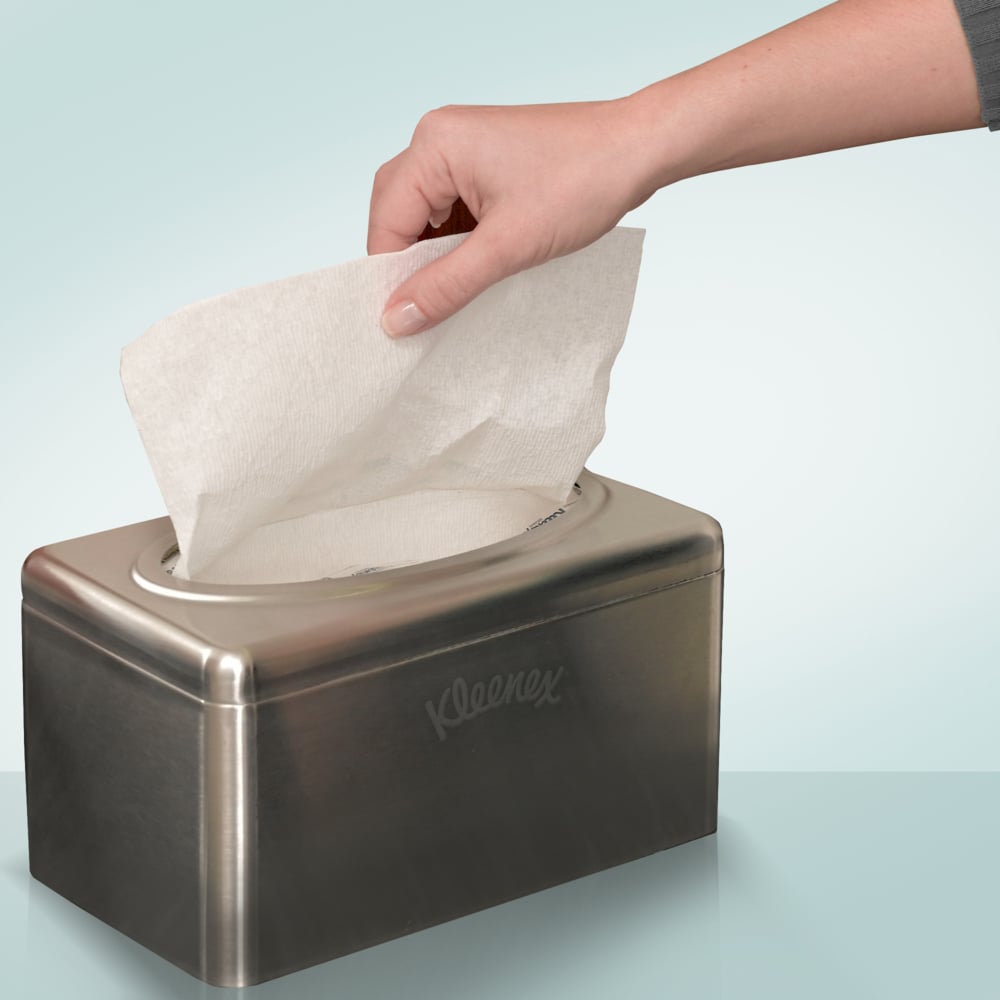 A hand pulls a tissue from a Kimberly-Clark GmbH Kleenex® towel dispenser made of stainless steel - pull-box/small, chrome-plated (2 dispenser boxes/carton), which stands on a light blue surface.