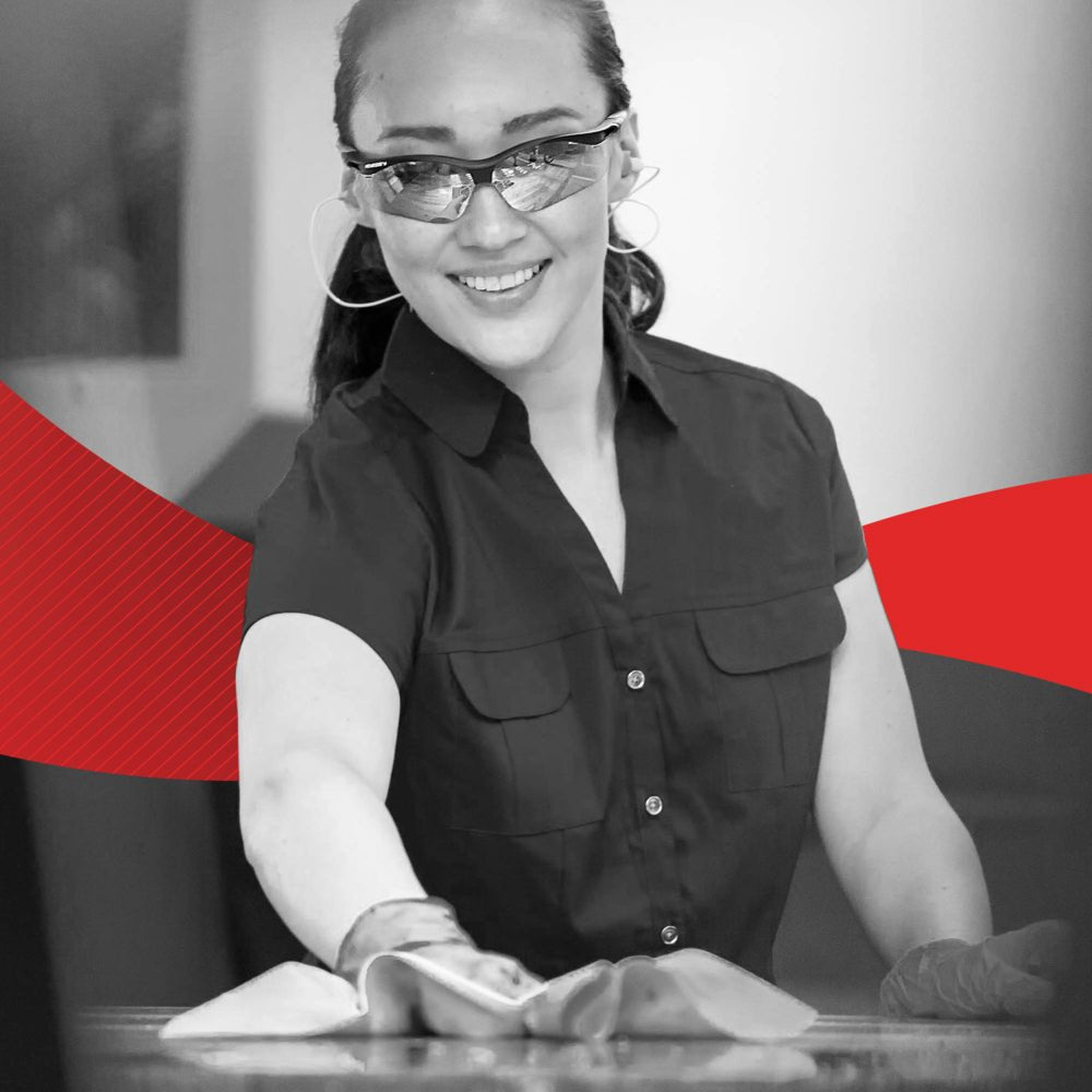 A woman with gloves and glasses smiles while cleaning a surface with a WypAll® X60 General Clean - BRAG™ Box cloth (41.80 × 28.30 cm) from Kimberly-Clark GmbH; a red graphic design appears in the background.