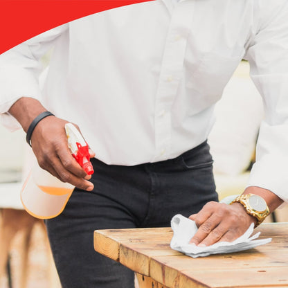 A person in a white shirt and black pants cleans a wooden table with WypAll® X60 General Clean cloths from Kimberly-Clark GmbH and holds a spray bottle. The scene shows efficient cleaning and a red curved edge at the top.