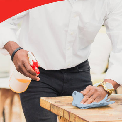 A person in a white shirt and watch cleans a wooden table with a blue cloth, possibly WypAll® L20 General Clean - BRAG™ Box from Kimberly-Clark GmbH. The image has a red curved edge at the top, emphasizing attention to detail.
