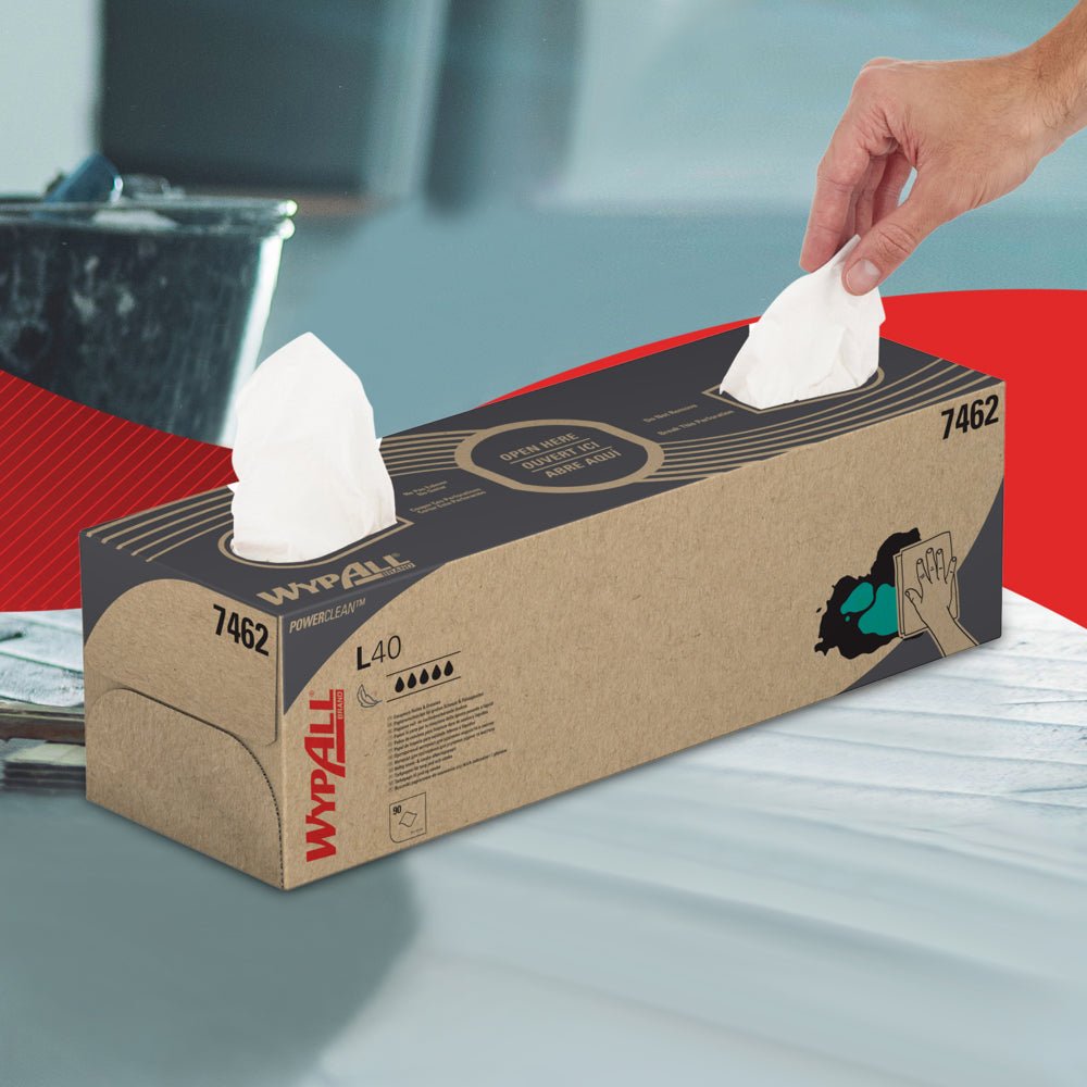 A hand pulls a WypAll® L40 PowerClean wipe from a rectangular white pop-up box by Kimberly-Clark GmbH, which is placed on a surface where another sheet can be seen; the box is adorned with black and red text and illustrations.