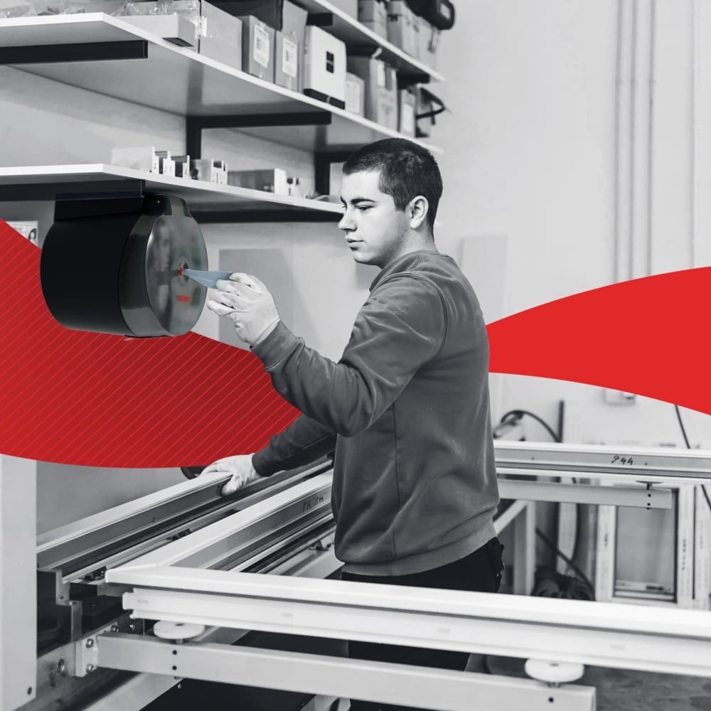 In a workshop, a person operates a wall-mounted device near shelves with WypAll® L10 paper wipes for food & hygiene centrefeed (6 rolls) by Kimberly-Clark GmbH; above it are boxes and a red swoosh accents the background.