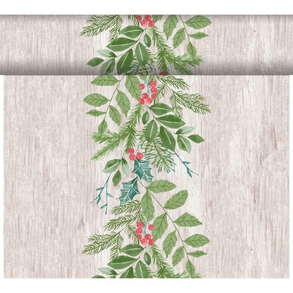 A vertical arrangement of green leaves, pine branches, and red berries decorates the Dunicel-Tete a Tete 0.4x24m table runner from Duni GmbH against a light wooden background with a festive repeat pattern at the top edge.