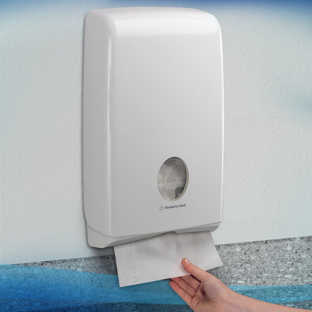 A hand pulls a Scott® SLIMFOLD towel from a white Kimberly-Clark GmbH wall dispenser with a viewing window showing folded towels inside. The hygienic dispenser is mounted on a light wall; it holds 16 clips x 110 sheets per carton.